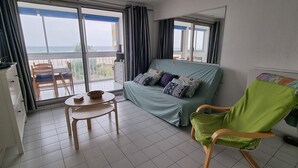 A balcony with a view of the sea, a sofa with pillows, a table with a bowl, and a chair with a blanket. - Appartement 2 Pièces Face à la mer (Valras-Plage)