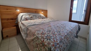 A bedroom with a wooden headboard, a patterned bedspread, a nightstand, and a window with a view. - Appartement 2 Pièces Face à la mer (Valras-Plage)