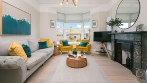 A modern living room with a sofa, armchairs, a coffee table, a fireplace, and a large window offering a view of the outside. - Summer Salt – St Ives – Sleeps 4 – Parking – Outside Space (St Ives)