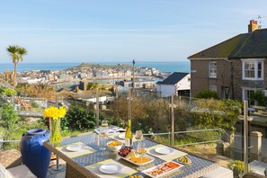 A table set for dining with a view of a coastal town, a large vase with flowers, and a bottle of wine. - Summer Salt – St Ives – Sleeps 4 – Parking – Outside Space (St Ives)