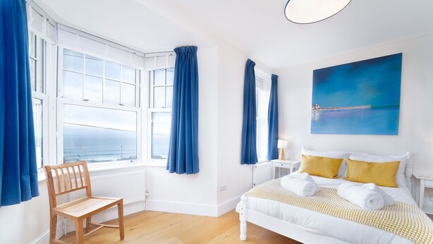 Une chambre à coucher avec un lit, une chaise, une fenêtre avec des rideaux, un tableau et une vue sur la mer.