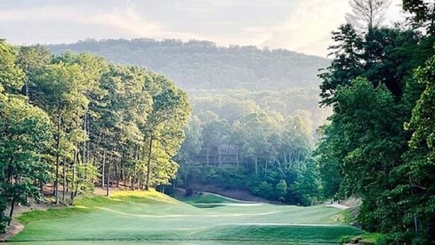 Ein Golfplatz mit einem Teich, umgeben von dichten Bäumen und Hügeln im Hintergrund.