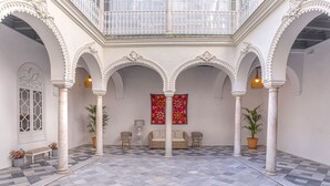 Une cour spacieuse dotée d’un sol à carreaux, de colonnes blanches, d’un balcon orné de balustrades travaillées et d’une tapisserie rouge à motifs.