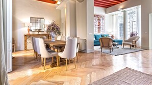 Une salle à manger spacieuse avec un parquet en bois disposé en chevrons, une grande table en bois et des chaises modernes. On y trouve une grande fenêtre ornée de vitraux, un canapé bleu et un vase décoratif rempli de fleurs.