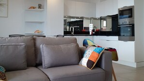 A modern living room with a grey sofa, a coffee table with a decorative mat, and a kitchen area with white cabinets and a built-in oven. - Porticcio, Superb air-conditioned T2, panoramic view of the Gulf of Ajaccio (Albitreccia)