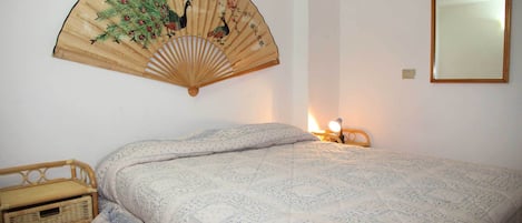 A bedroom with a bed, a wall-mounted fan, a bedside table, and a mirror.
