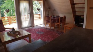 A cozy living room with a wooden floor, a red patterned rug, a dining area with a table and chairs, and a large window with a view of greenery. - Gemütliches Ferienhaus am Twistesee im Ferienland Waldeck-frankenberg (Bad Arolsen-Wetterburg)