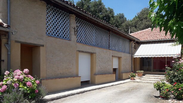 Un bâtiment à la façade beige, doté de fenêtres à croisillons et d’un toit recouvert de tuiles rouges. On y voit un escalier menant à une porte, ainsi que des plantes en pot.
