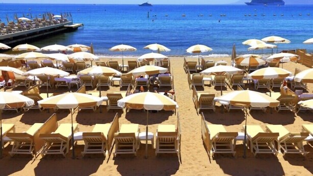 Une plage bordée de rangées de chaises longues et de parasols, une jetée s’avançant dans la mer, et plusieurs bateaux en arrière-plan.