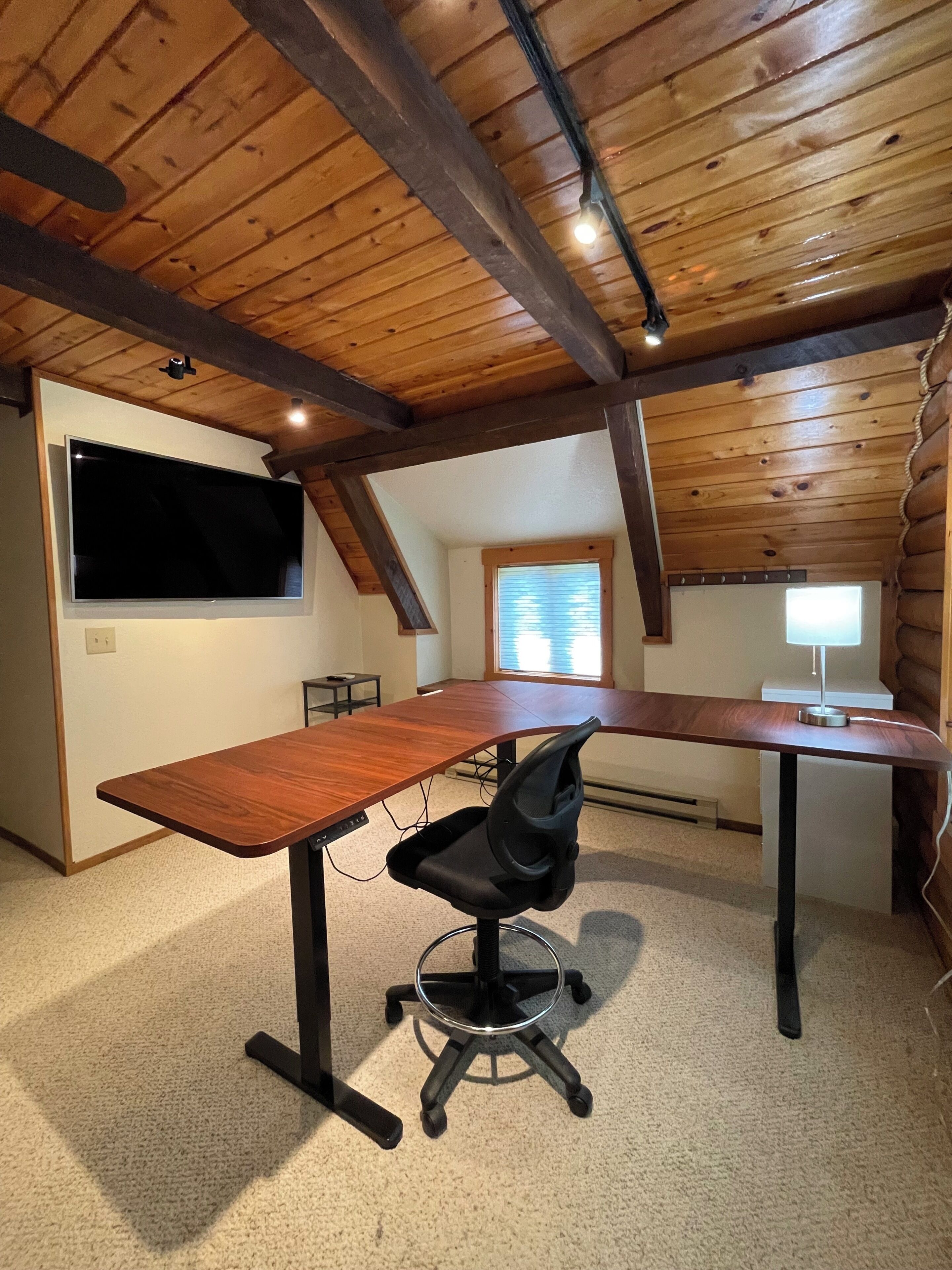 Een kamer met houten plafond, een groot bureau, een zwarte bureaustoel, een aan de muur bevestigde televisie en een raam met jaloezieën.