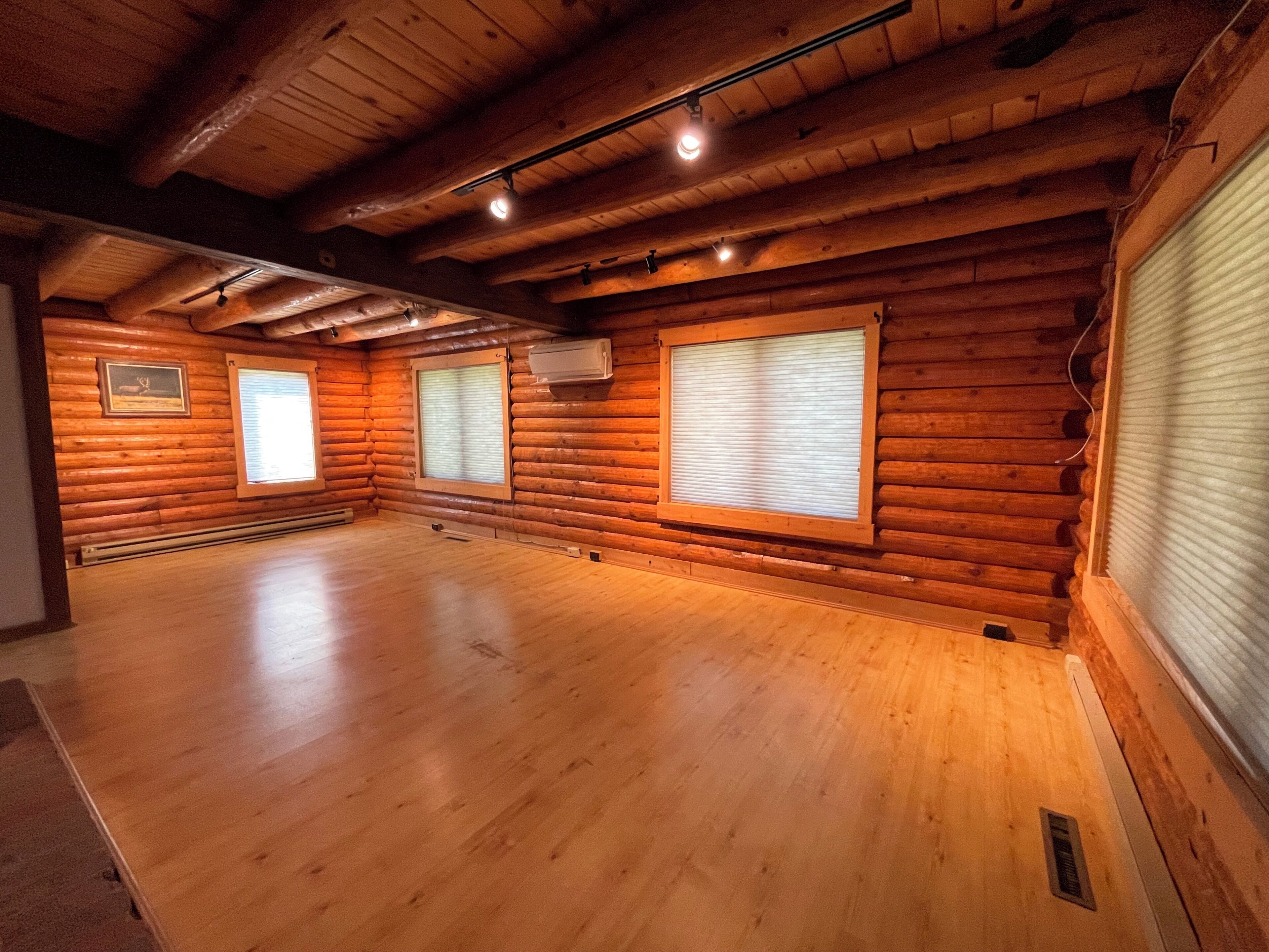 A log cabin interior with wooden walls, a wooden floor, and a large window with blinds.