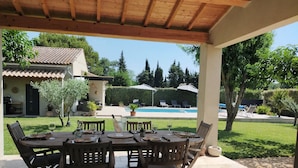 A covered patio with a wooden table and chairs, a pool in the background, and a house with a tiled roof. - Lou Mazet provençal à Eygalières - Private swimming pool (EYGALIERES)