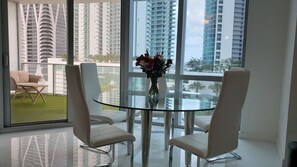 A modern dining area with a glass table and white chairs, a vase with flowers, and a view of tall buildings through large windows. - Luxury Apartment (Miami)