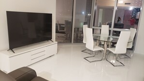 Una sala moderna con un televisor de pantalla plana sobre un mueble blanco, una mesa de comedor con superficie de vidrio y sillas blancas, y un espejo grande que refleja la habitación.