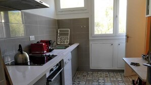 Une cuisine de taille réduite, équipée d’une cuisinière à gaz, d’un grille-pain rouge et d’une bouilloire. Le sol est carrelé et présente un motif floral. Une fenêtre offre une vue sur les arbres.