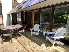 An outdoor deck with a round table and chairs, a white Adirondack chair, and a glass door leading to a patio area. - 2026 Seasonal Summer Rental (Winhall)
