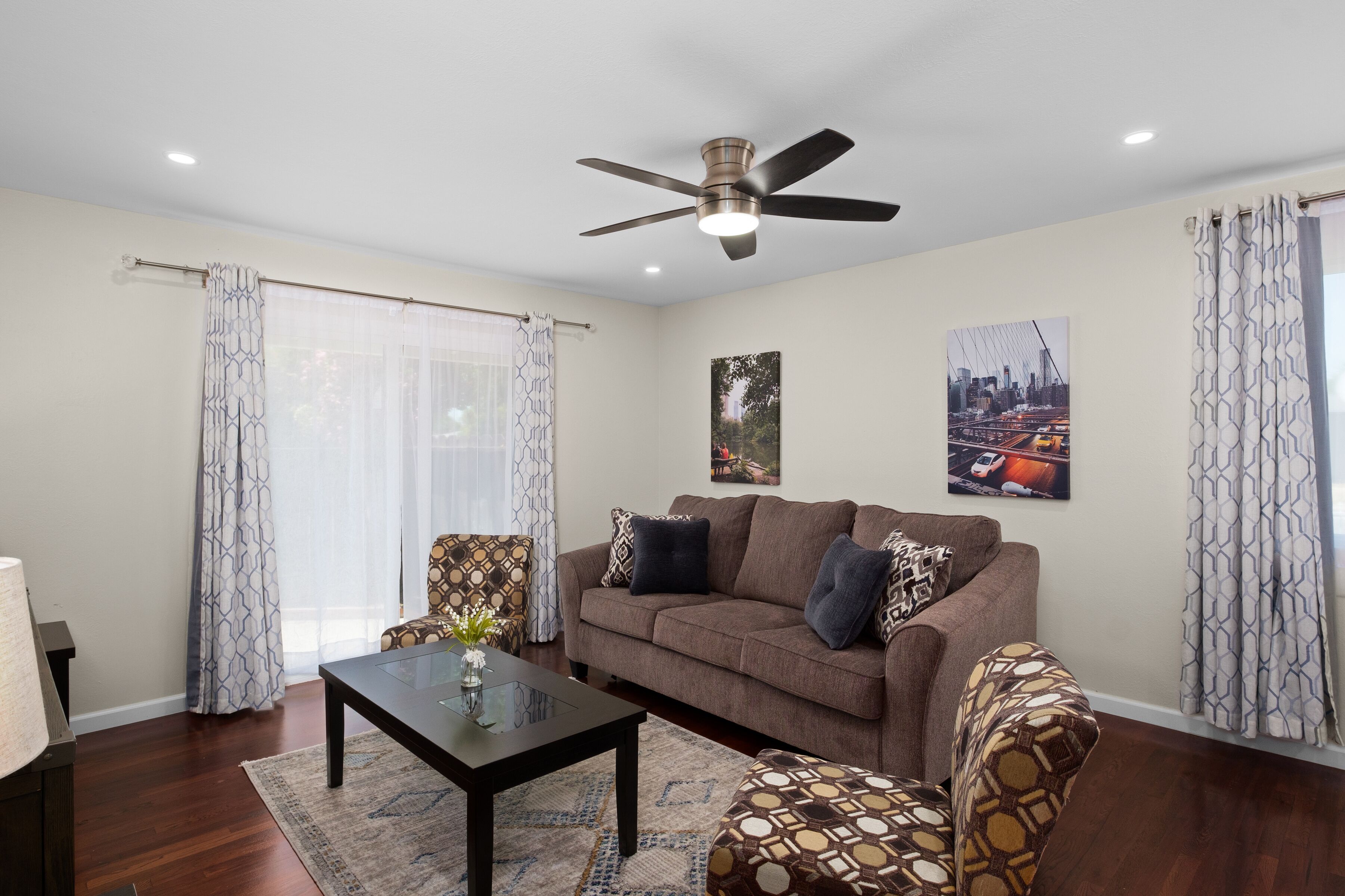 A living room with a brown sofa, patterned armchairs, a glass coffee table, and a ceiling fan.