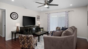 A living room with a flat-screen TV, a ceiling fan, a clock, and a sofa.