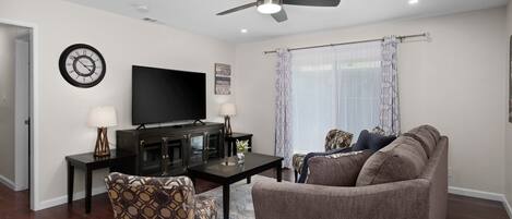 A living room with a flat-screen TV, a ceiling fan, a clock, and a sofa.
