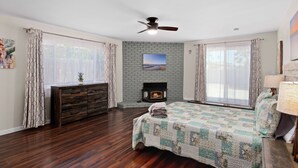 A bedroom with a bed, a fireplace, a ceiling fan, and large windows.
