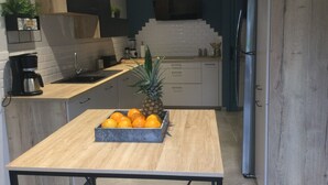 Eine moderne Küche mit einem Holztisch, einer Obstschale mit Orangen und einer Ananas, einer Kaffeemaschine und einem Kühlschrank.