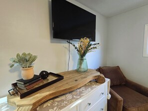 Un salon avec un téléviseur à écran plat fixé au mur, une table basse en bois sur laquelle se trouve un plateau avec des livres et une plante, un vase rempli de fleurs et un canapé marron.