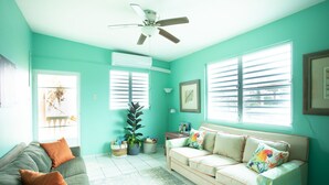 A living room with teal walls, a ceiling fan, two windows with blinds, a gray sofa with orange cushions, a beige sofa with colorful pillows, and a small table with a lamp.