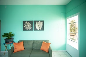 Een kamer met teal muren, een groene bank met oranje kussens, een potplant en twee kunstwerken met schelpen in lijsten.
