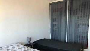 A small, single-bed room with a bed covered in a polka dot blanket, a bedside table with a lamp, and a window with sheer curtains. - T2 , 50m² Avec vue mer à Carnon Plage (Mauguio)
