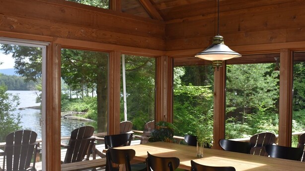 A wooden interior with a dining table and chairs, large windows, and a view of a lake. - Luxurious Waterfront with Mountain Views (Tupper Lake)