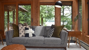 A cozy living room with a grey sofa, various patterned cushions, a wooden side table, and a large window offering a view of trees and a lake. - Luxurious Waterfront with Mountain Views (Tupper Lake)