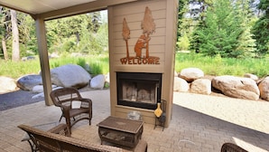 A covered outdoor seating area with wicker furniture, a fireplace with a 'WELCOME' sign, and a natural backdrop of trees and rocks. - Hart of McCall - Mountain retreat with hot tub and sauna near downtown and golf (McCall)