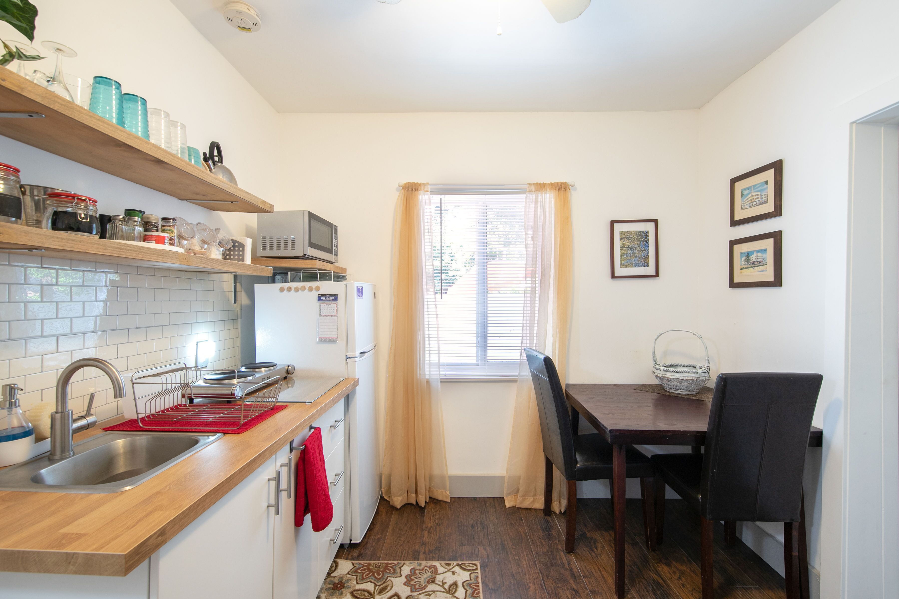 A compact kitchen with a sink, microwave, and wooden shelves. There is a dining table with chairs and a window with curtains.