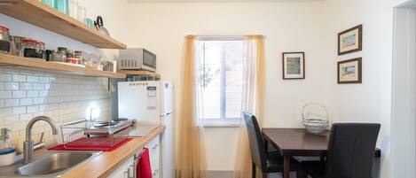 A compact kitchen with a sink, microwave, and wooden shelves. There is a dining table with chairs and a window with curtains.