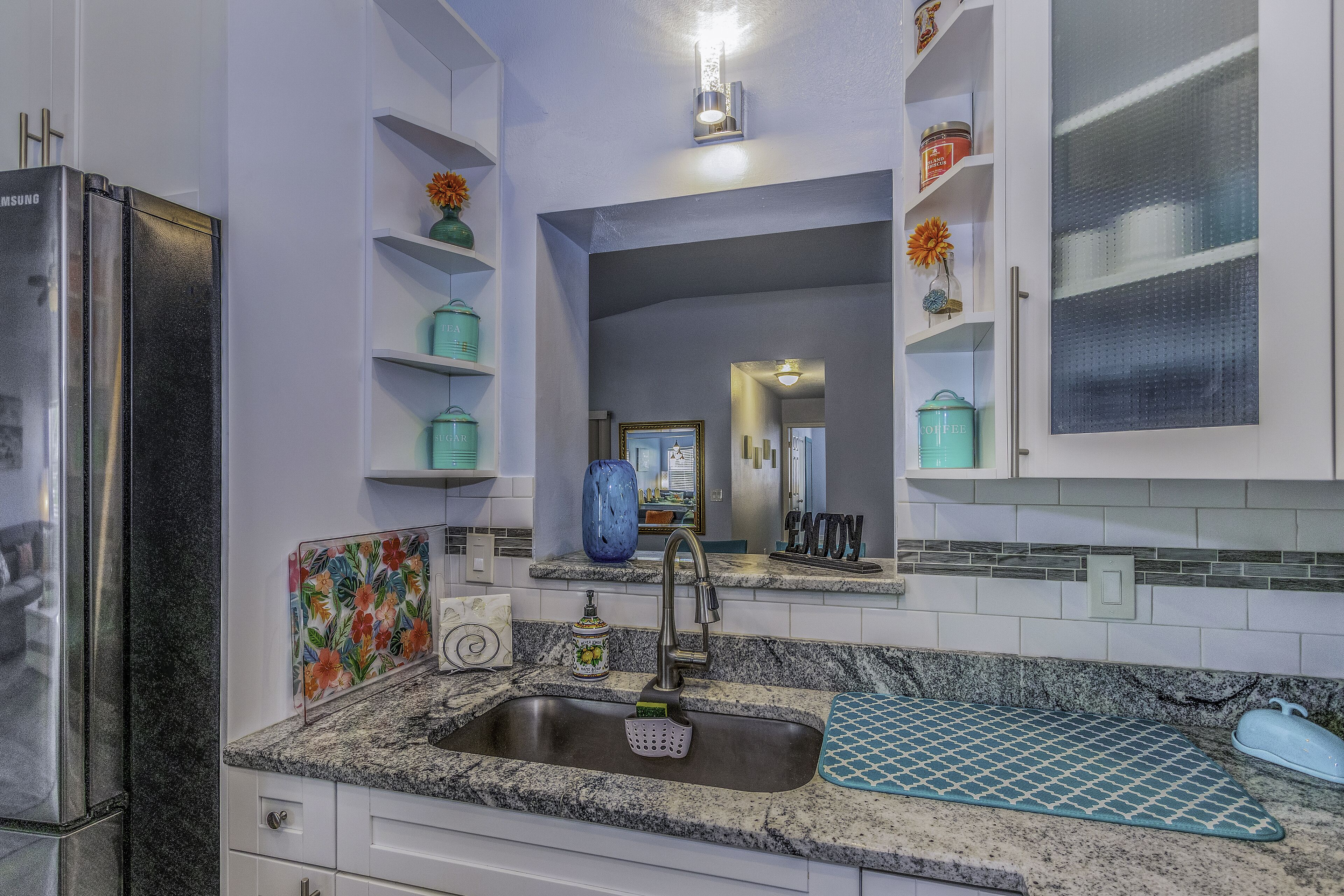 Una cocina moderna con un refrigerador de acero inoxidable, encimeras de granito, un lavamanos con grifo y objetos decorativos en estantes.