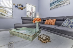 A living room with a grey sofa, a glass coffee table with a vase of flowers, and decorative plates on the wall. - Beautiful, spacious, 4 king beds home with heated and covered pool (Clearwater)