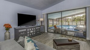 Una sala de estar moderna con un televisor de pantalla plana, sofá gris y vista a una piscina exterior a través de amplias puertas de cristal.