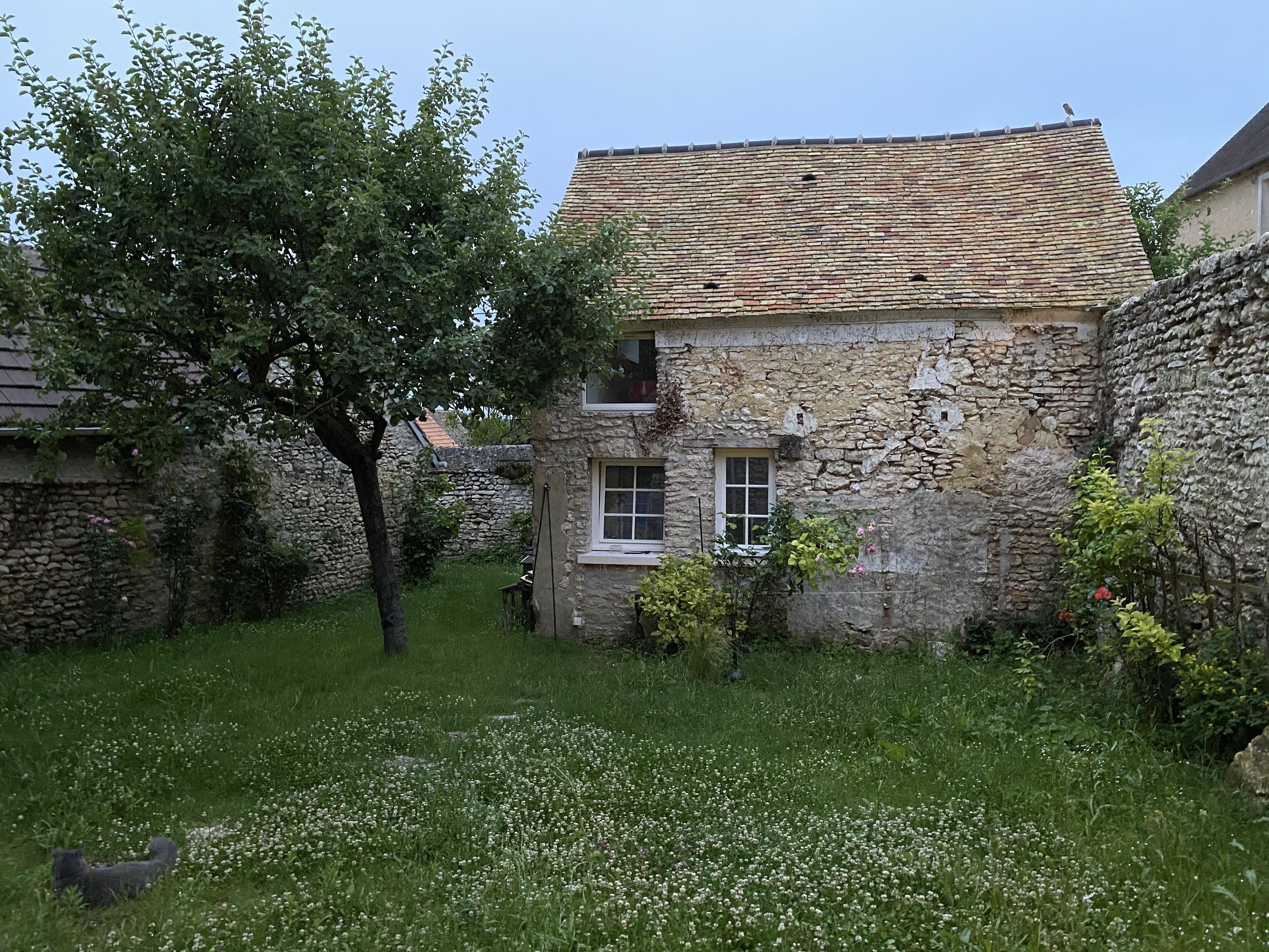 A stone building with a tiled roof, a small window, and a tree in the foreground.