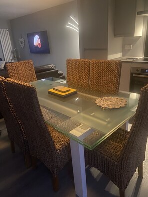 A modern dining area with a glass table surrounded by woven chairs, a television mounted on the wall, and a kitchen in the background.