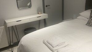 A neatly made bed with folded white towels, a white bedside table with a mirror, a small round mirror, and a grey door.