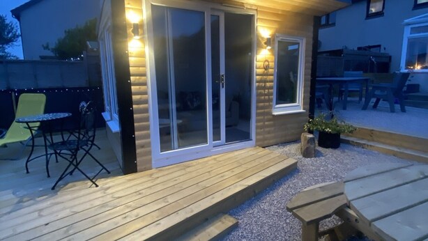A small wooden structure with a deck, a sliding glass door, and a patio area with benches.