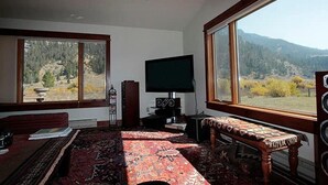 A living room with a large window, a flat-screen TV, a bench, and a speaker system. - Private Island in Big Sky-Great Fishing & Hunting (Big Sky)