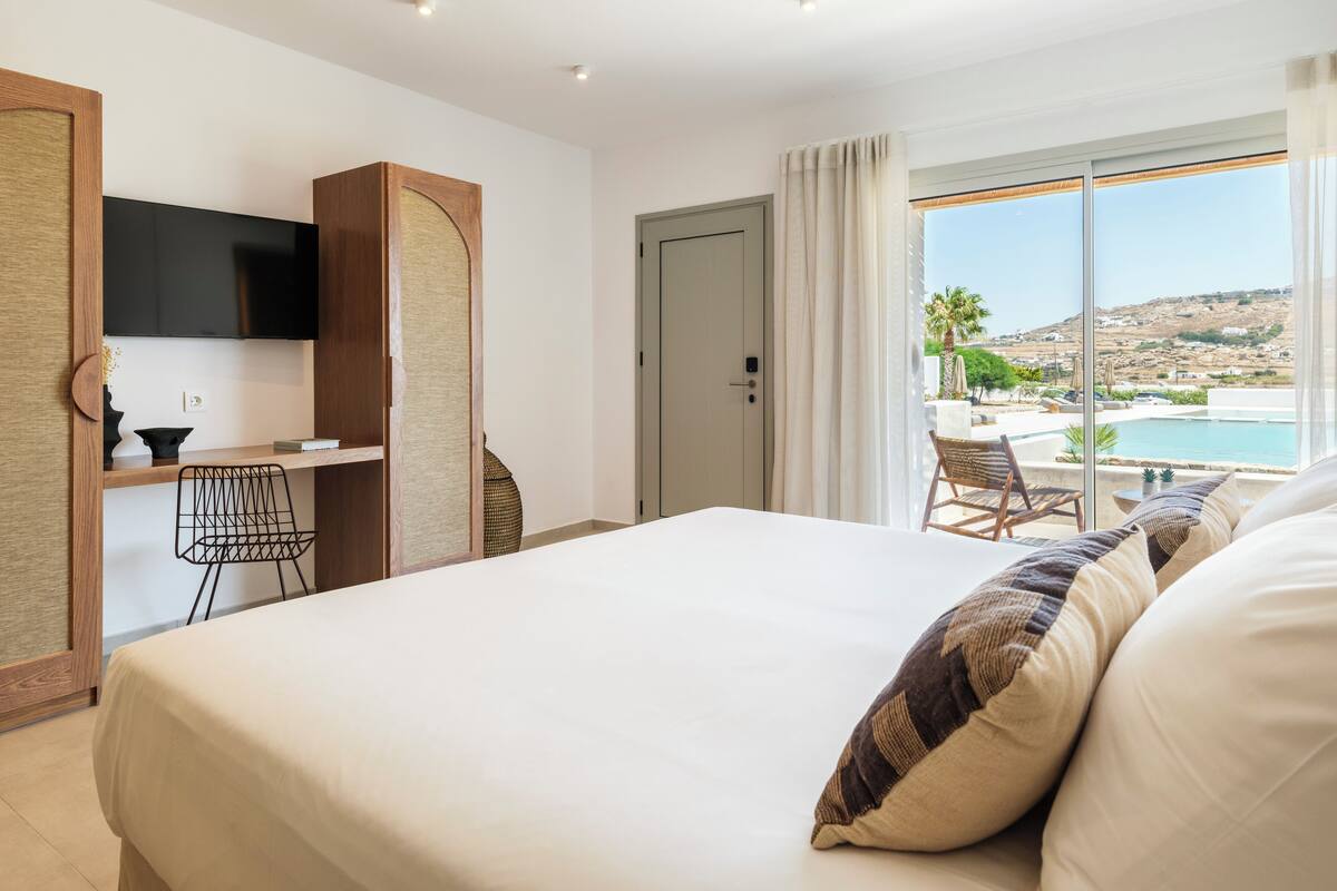 A hotel room with a bed, a desk, a chair, a TV, and a view of the sea.
