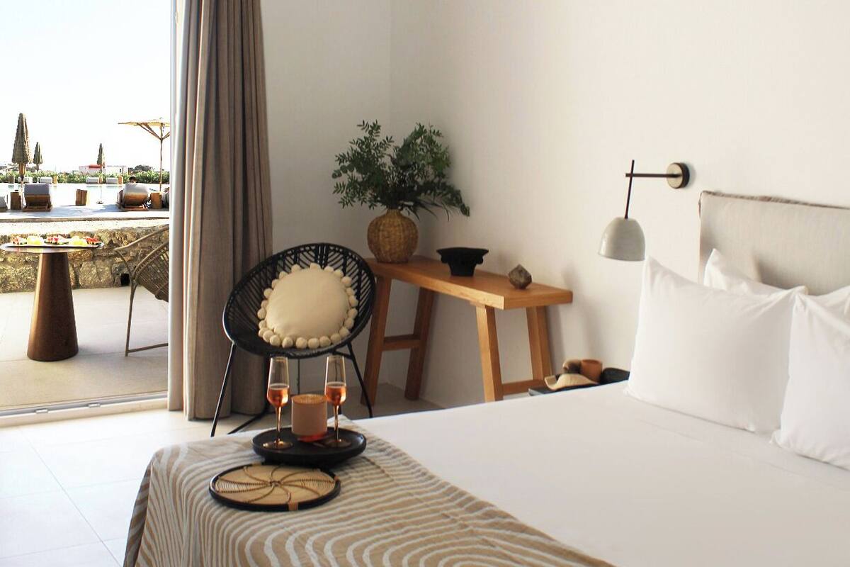 A modern hotel room with a large bed, a small table, a chair, and a view of an outdoor pool.