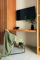 Quarto com escrivaninha de madeira, TV preta fixada na parede, planta em vaso e manta verde sobre uma cadeira.