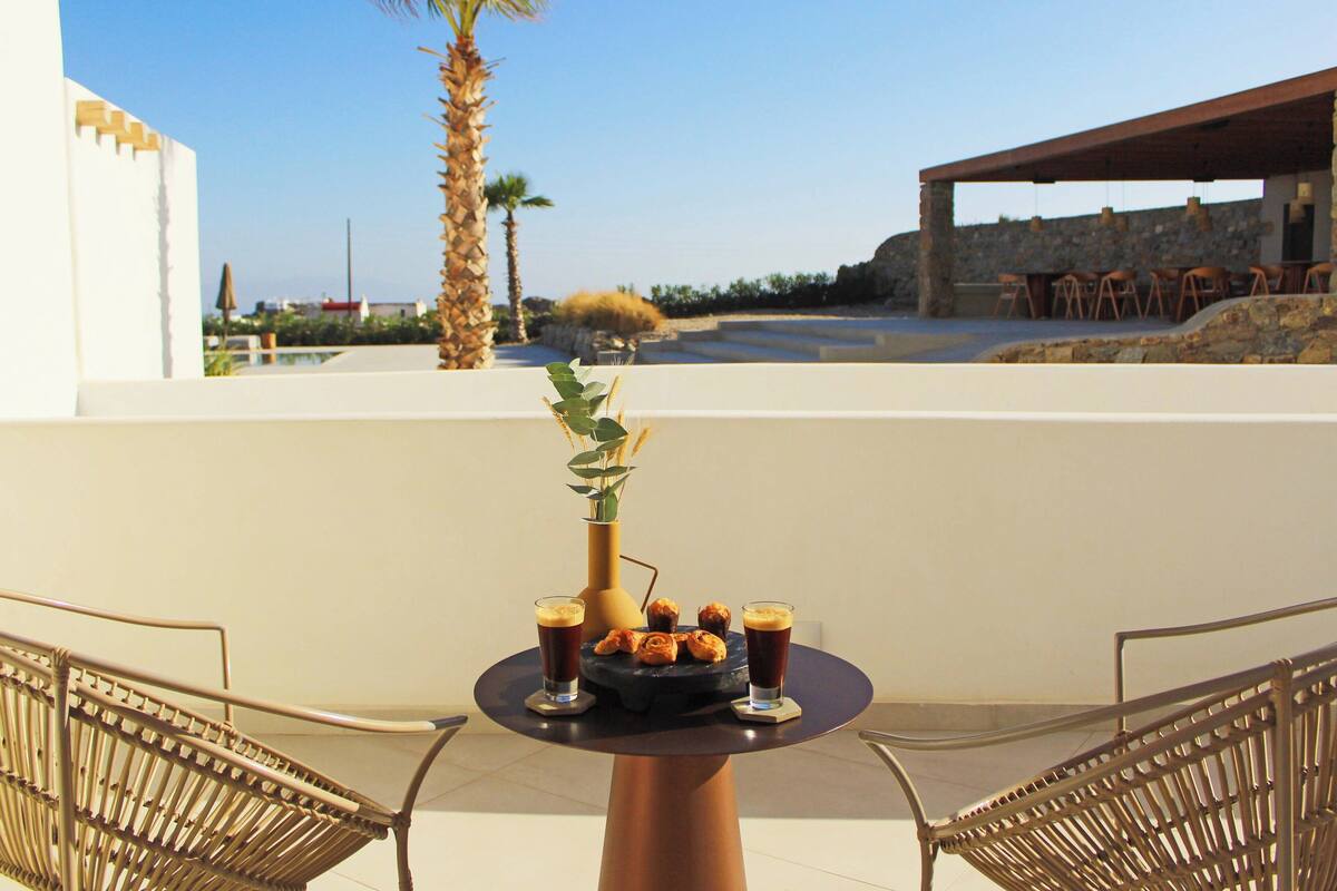 A table with drinks and pastries, a vase with greenery, and a view of a pool and palm trees.