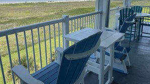 Un espace extérieur abrité, aménagé avec des chaises et des tables blanches et bleues, donnant sur une plage verdoyante avec l’océan à l’horizon.