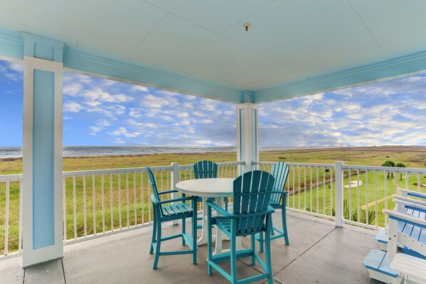 A covered patio with a table and chairs, overlooking a grassy field and ocean. - Pointe West Top Floor Renovated Spacious Beachfront 3/3 Resort Condo (Galveston)