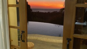 Une vue à travers une porte en bois donnant sur une piscine, avec un coucher de soleil en arrière-plan.