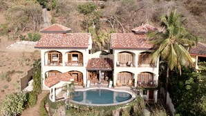 Une grande maison à deux étages avec une piscine, entourée de palmiers et d’une végétation aride.
